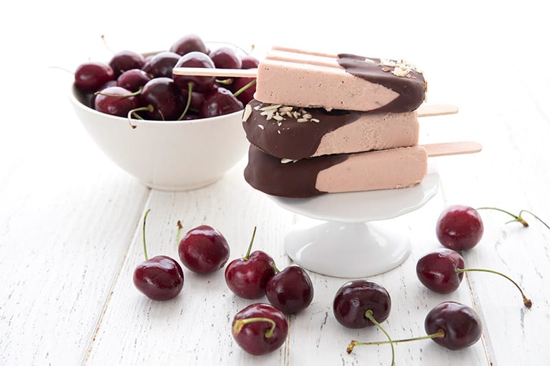 A stack of three cherry popsicles surround by cherries and a bowl of cherries in the background