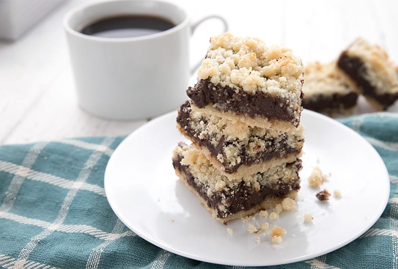 A white plate with a stack of fudge crumb bars over a teal plaid napkin, with a cup of coffee in the background
