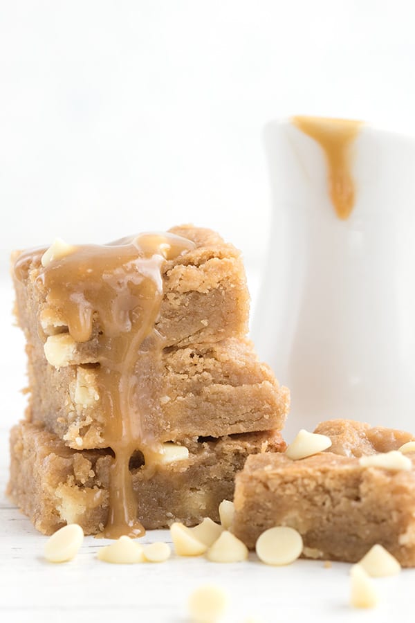 Keto white chocolate blondies in a stack with caramel sauce dripping down, with the pitcher of caramel sauce in behind.