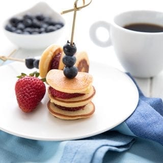Two mini pancake skewers on a white plate with a strawberry. A cup of coffee and a bowl of blueberries in the background.
