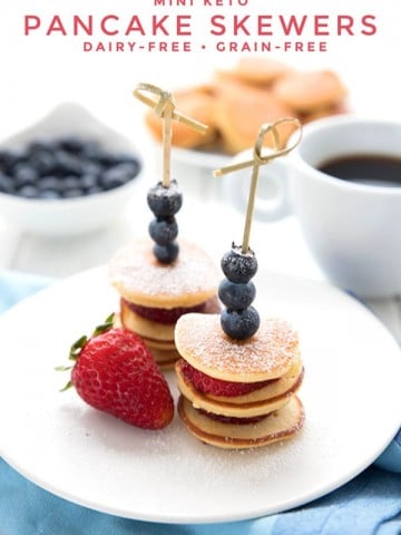 Titled image of keto mini pancake skewers on a white plate over a blue napkin. A cup of coffee, a bowl of blueberries and a plate of mini pancakes in the background.
