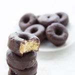A stack of keto chocolate covered mini donuts with a bite taken out of the top one. More mini donuts in the background on a white plate