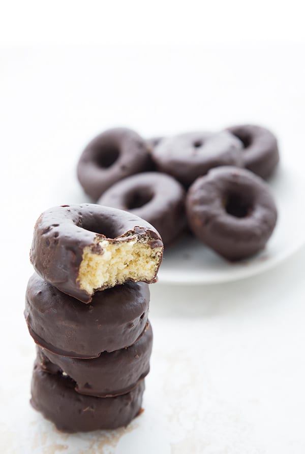 A stack of keto chocolate covered mini donuts with a bite taken out of the top one. More mini donuts in the background on a white plate