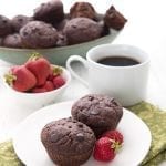 Two double chocolate zucchini muffins sit on a white plate over a green patterned napkin. A cup of coffee, a bowl of berries, and a bowl of muffins in the background.