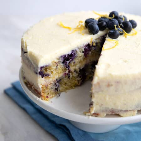 Keto Lemon Blueberry Cake with a slice cut out of it, on a white cake stand over a blue napkin.