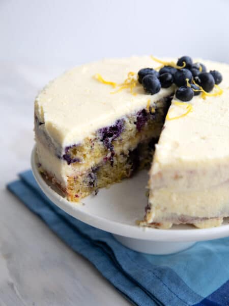 Keto Lemon Blueberry Cake with a slice cut out of it, on a white cake stand over a blue napkin.