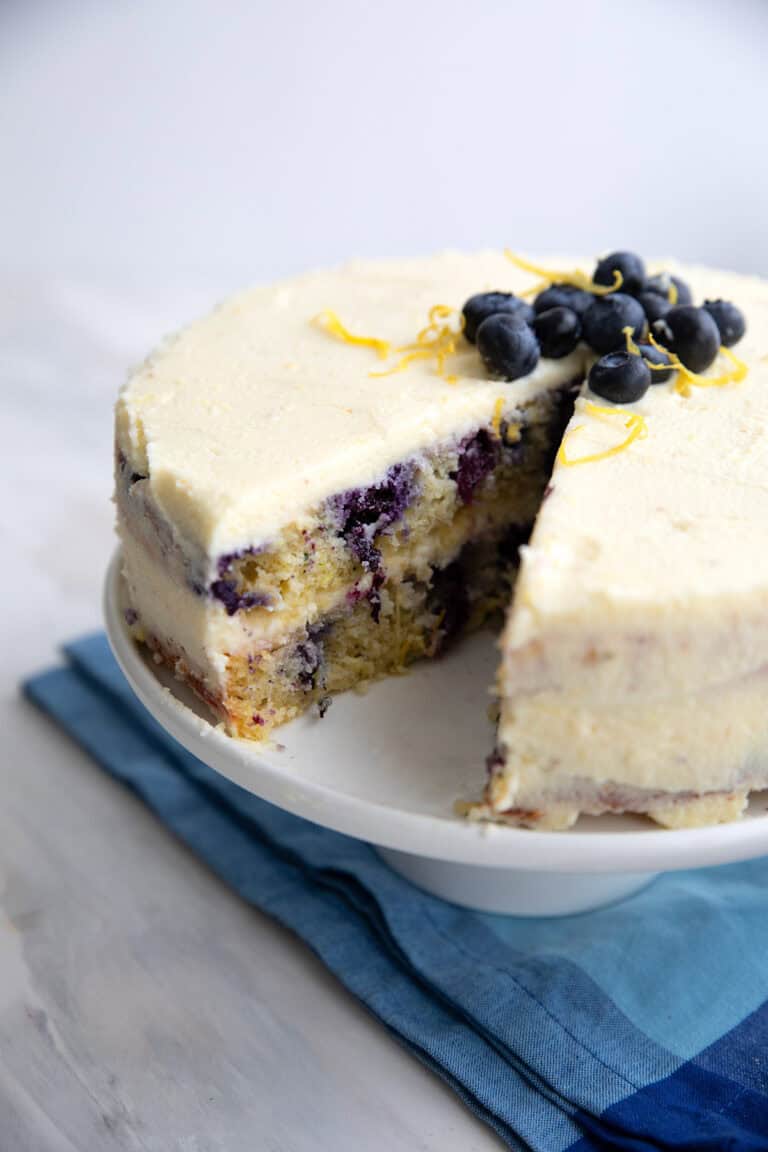 Keto Lemon Blueberry Cake with a slice cut out of it, on a white cake stand over a blue napkin.