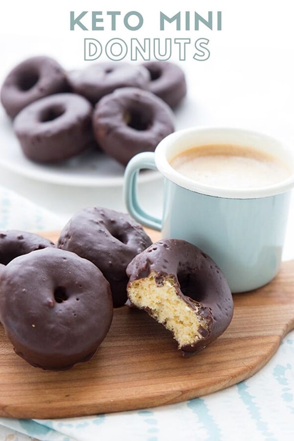 Titled image: a small cutting board with a cup of coffee and some chocolate covered mini donuts. A bite is taken out of the front donut. There is a white plate in the background with more donuts.
