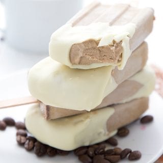 Close up shot of keto coffee popsicles stacked on a white plate with coffee beans. A bite has been taken out of one popsicle