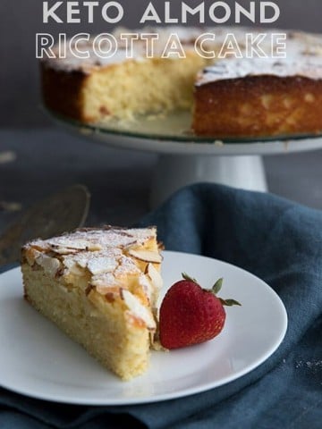 Titled image of keto almond ricotta cake. A slice of cake on a white plate with a strawberry, the rest of the cake on a stand in the background.
