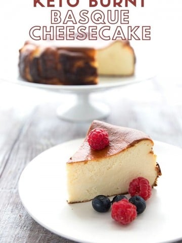 Titled image of Keto Burnt Basque Cheesecake. A slice on a white plate with berries, with the rest of the cake in the background on a cake stand.