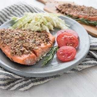 A grey plate with pecan crusted salmon, coleslaw, and fresh sliced tomatoes sits on a grey and white striped napkin. A cutting board with another piece of salmon in the background.