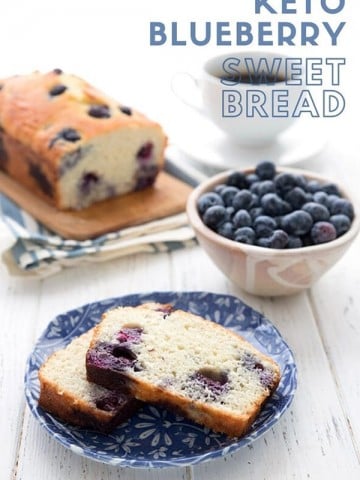 Titled image of keto blueberry coconut flour bread. Two slices on a blue patterned plate, in front of a bowl of blueberries, with the rest of the loaf in the background.