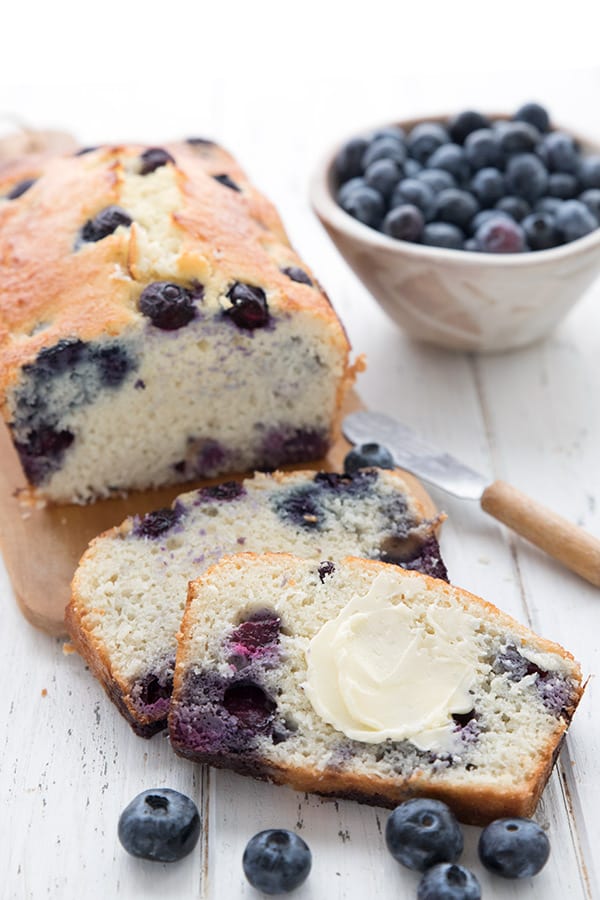 A slice of keto blueberry coconut bread with butter on it, in front of a cutting board with the loaf of bread and a bowl of blueberries