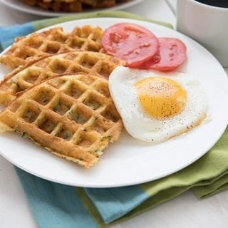 A white plate filled with eggs, zucchini waffles and tomatoes sites over a blue and green napkin