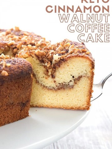 Titled image of Keto Cinnamon Walnut Coffee Cake, a slice being taken out of the large cake on a white cake stand.