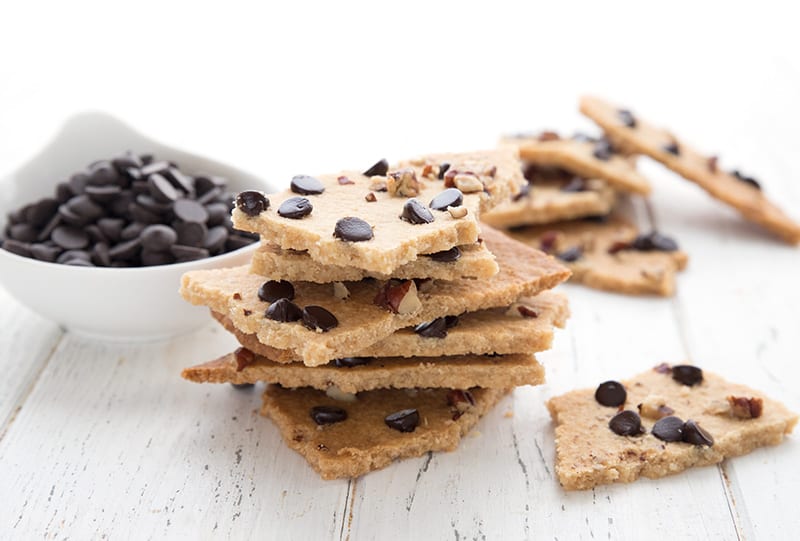 Keto cookie brittle piled around a white table with a bowl of chocolate chips.