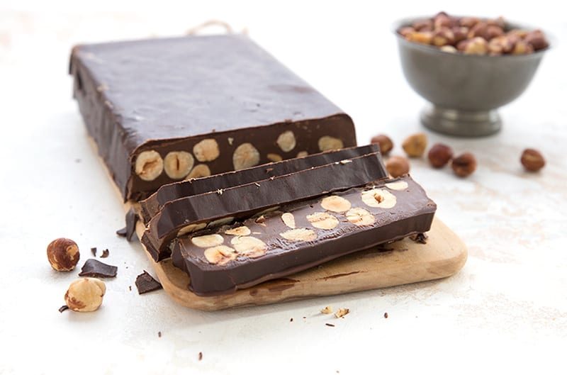 Keto chocolate hazelnut torrone cut into slices on a wooden cutting board, with a bowl of toasted hazelnuts in the background.