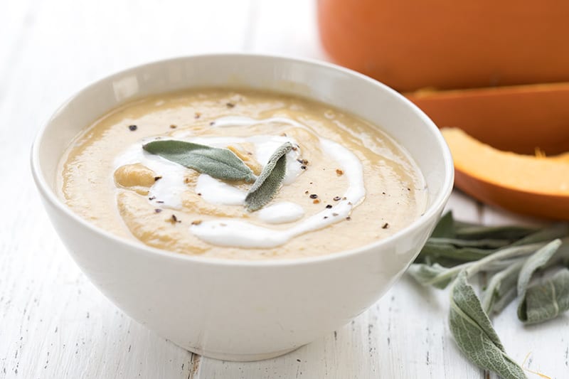 A white bowl filled with roasted pumpkin soup with sliced pumpkin and sage leaves in the background.