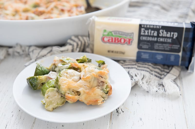 A white plate full of keto brussels sprouts casserole beside a brick of Cabot cheddar cheese.