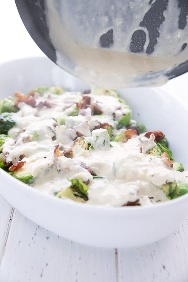 Creamy keto cheese sauce being poured over Brussels sprouts and bacon in a white casserole dish.