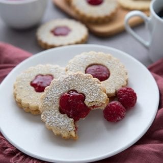 Three keto Linzer cookies on a white plate over a red napkin, with more cookies in the background and a bowl of the raspberry filling
