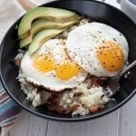 Cauliflower Bacon Grits in a black bowl, topped with fried eggs and avocado