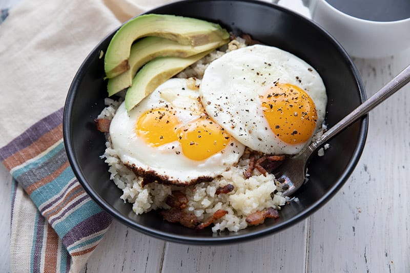 Cauliflower Bacon Grits in a black bowl, topped with fried eggs and avocado