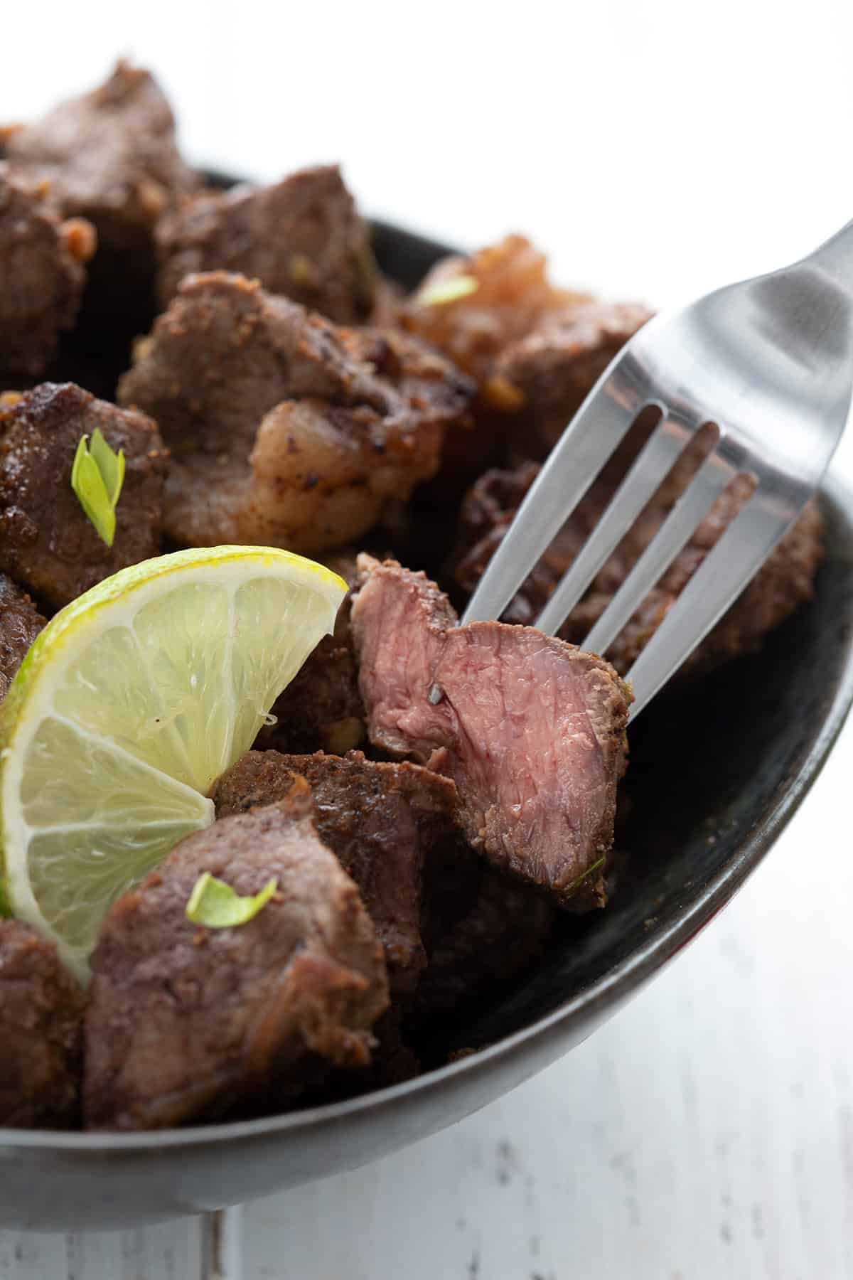 Close up shot of air fryer steak bites in a bowl, with a fork lifting one out. The bite is cut open to show the pink inside.