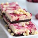 Keto Raspberry White Chocolate Cheesecake Bars stacked on a rectangular white tray with white chocolate chips sprinkled on top.