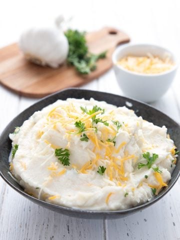 A black bowl full of cauliflower mashed potatoes in front of a bowl of cheese and a cutting board with garlic and parsley.