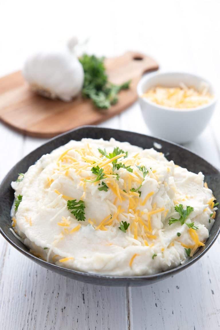 A black bowl full of cauliflower mashed potatoes in front of a bowl of cheese and a cutting board with garlic and parsley.