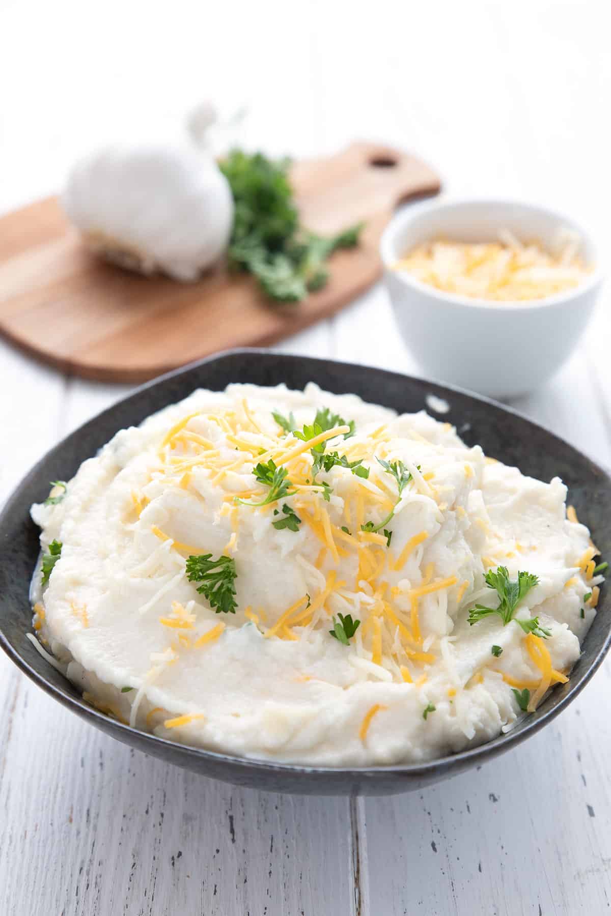 A black bowl full of cauliflower mashed potatoes in front of a bowl of cheese and a cutting board with garlic and parsley. 