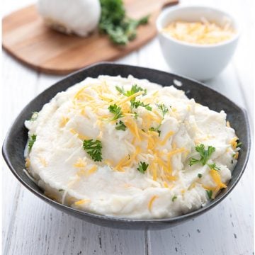 A black bowl filled with creamy mashed cauliflower topped with cheddar and parsley.