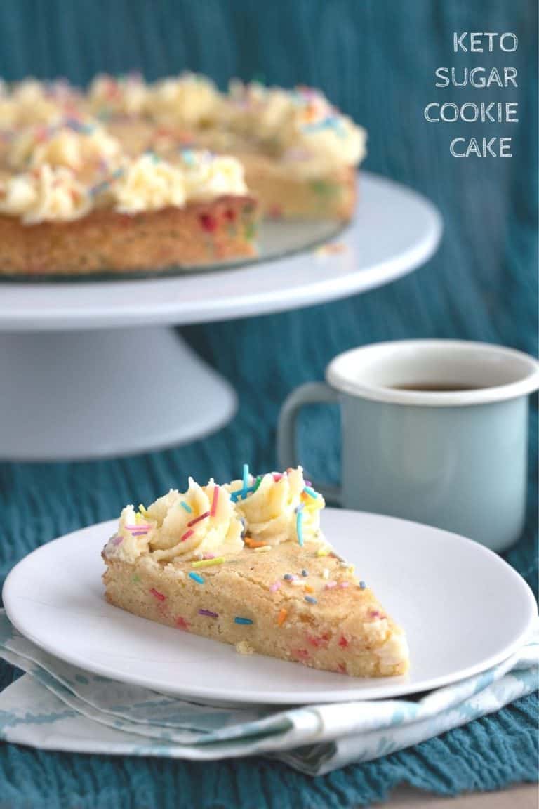 Titled image: a slice of keto sugar cookie cake on a white plate, with the rest of the cake in the background on a cake stand.