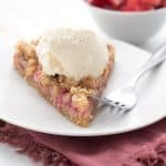 A slice of keto rhubarb crumble tart on a white plate with a scoop of sugar-free vanilla ice cream.
