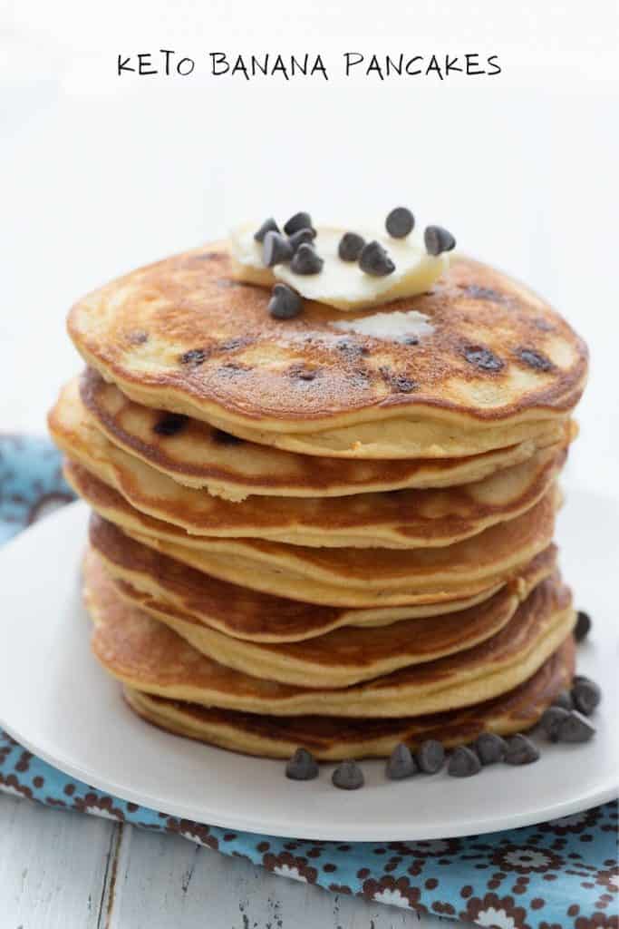 Titled image: A stack of keto banana pancakes on a white plate over a blue patterned napkin, with butter and chocolate chips on top.