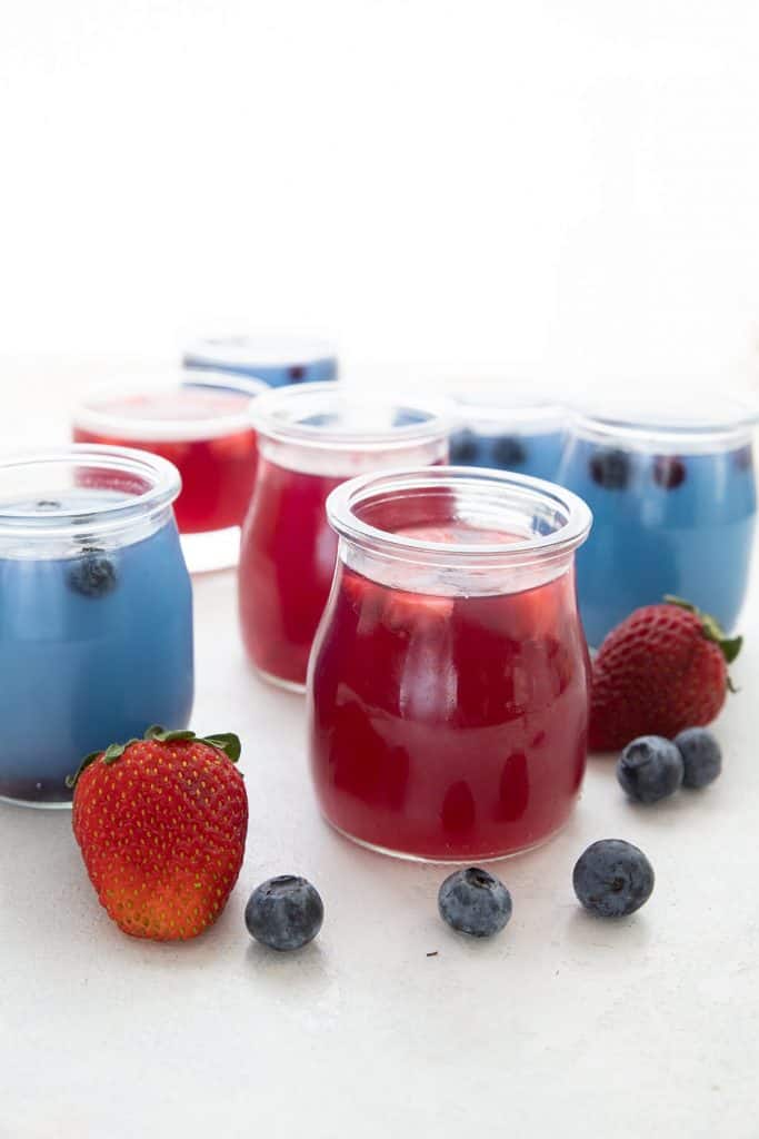 Small glass jars filled with keto sugar free jello, some blueberry and some strawberry, with fresh berries scattered around.