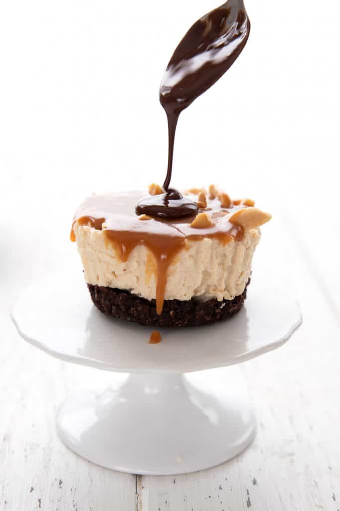 A spoon drizzling chocolate over a keto mini cheesecake on a small white cake stand.