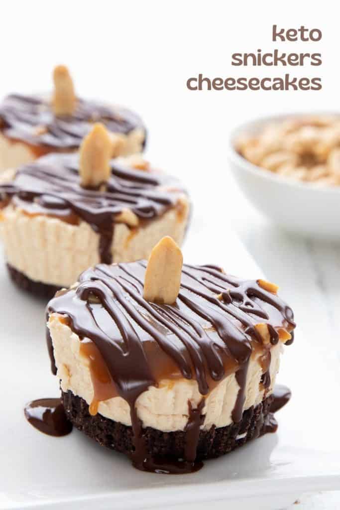 Titles image of three mini keto "Snickers" cheesecakes on a white tray, with a bowl of peanuts in the background.