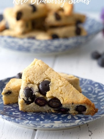Titled image of two keto blueberry scones an a blue patterned plate, with a larger plate of scones in the background.