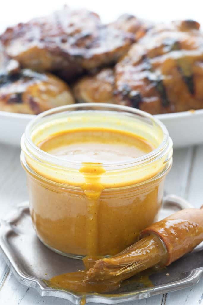 A jar full of Carolina Mustard Barbecue Sauce in front of a plate of grilled chicken.