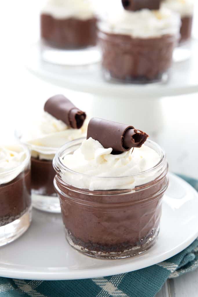 3 mini keto chocolate cream pies in jars, in front of a cake stand with more pies on it.
