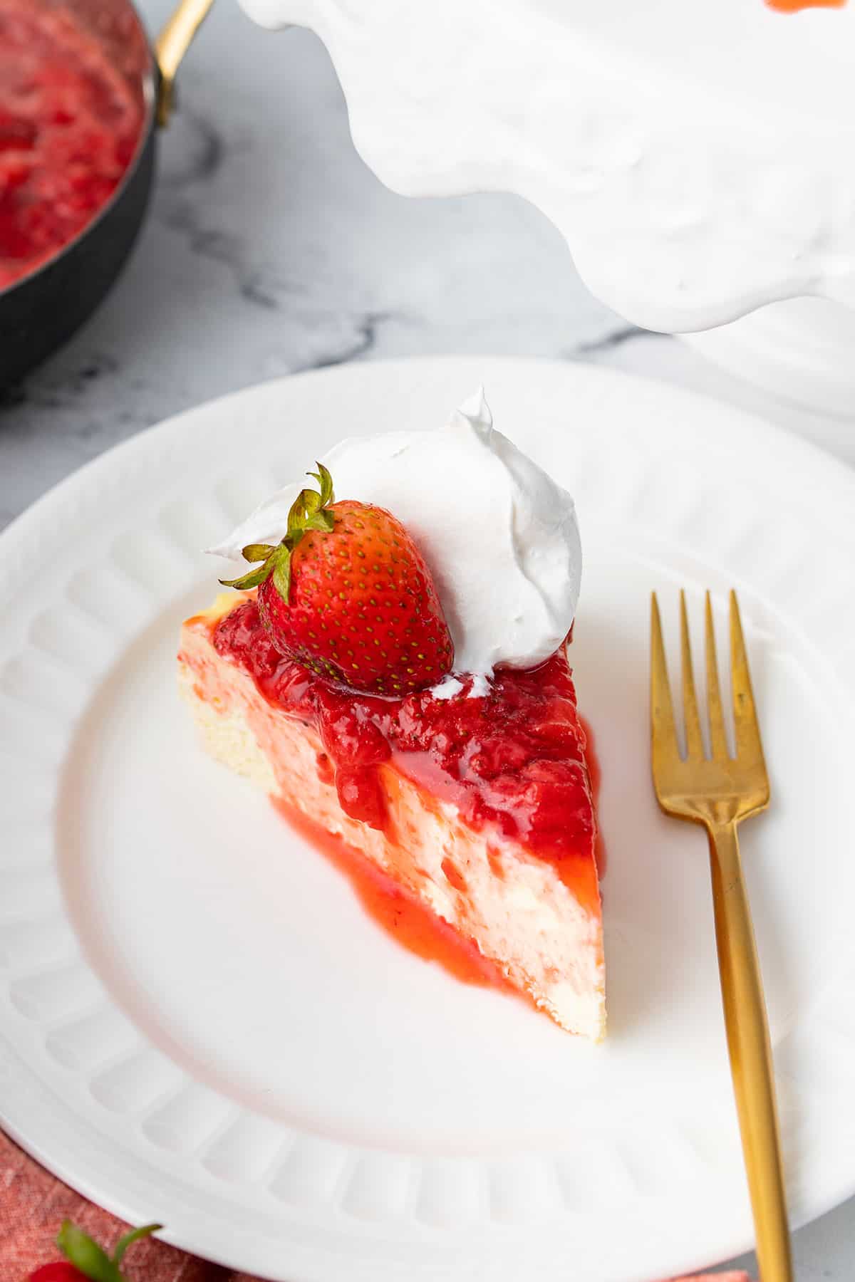 A slice of keto cheesecake on a white plate, with strawberry sauce and whipped cream on top. 