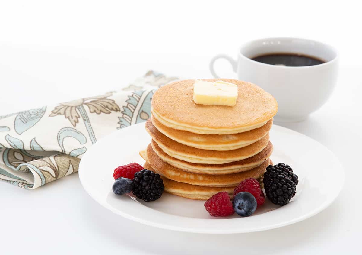A stack of keto pancakes on a white plate with a pat of butter on top. A cup of coffee and a napkin in the background.