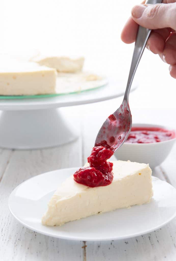 A slice of crustless keto cheesecake on a white plate with raspberry sauce being spooned over it.