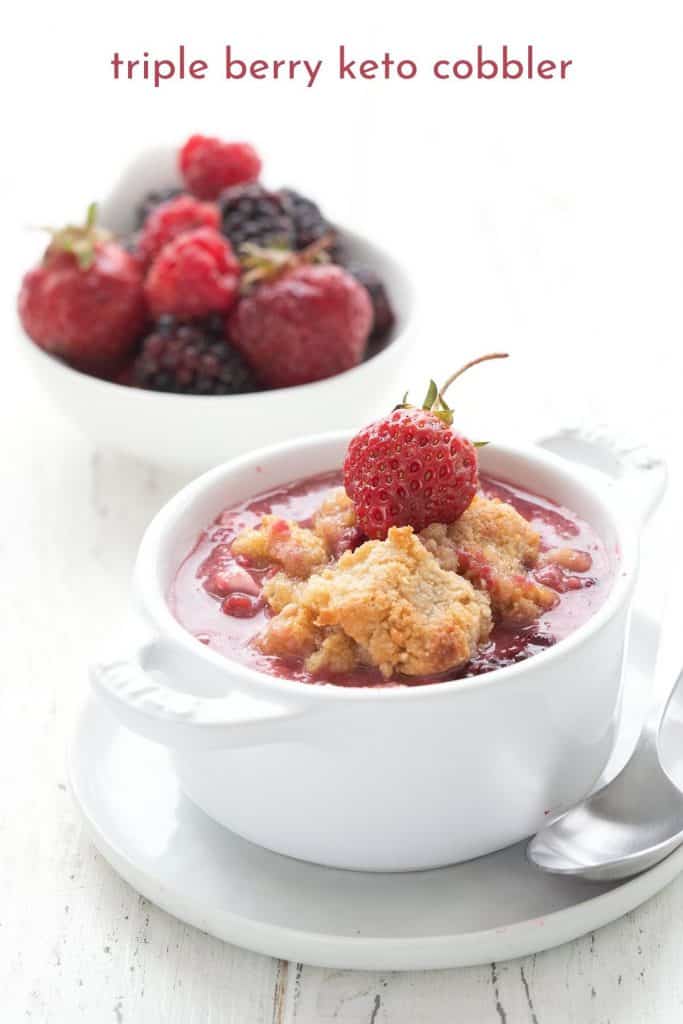 Titled image of triple berry keto cobbler in a white ramekin, with a bowl of berries in the background.