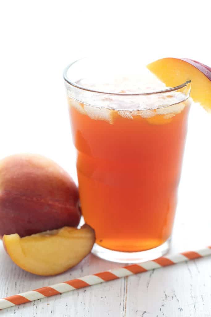 A glass full of keto sweet tea with peaches and a striped orange straw, on a white wooden table.