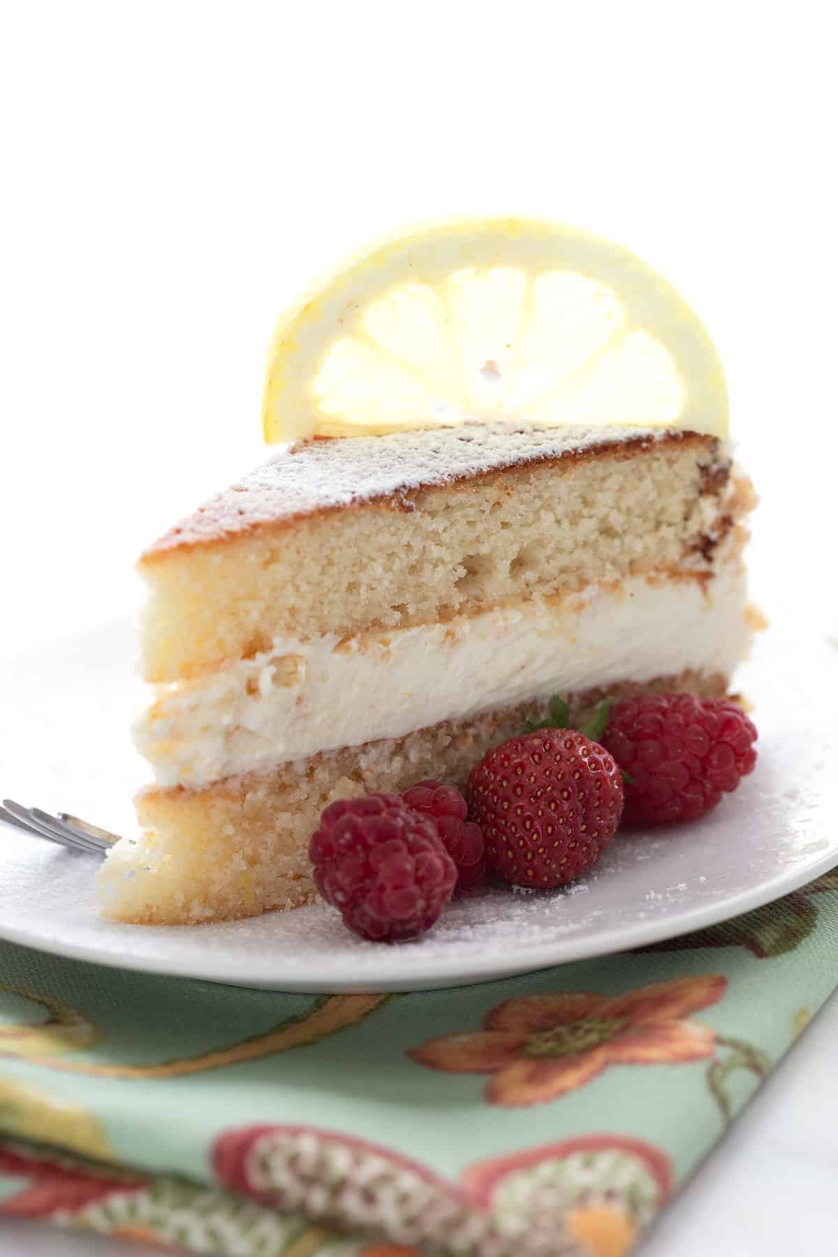 A slice of keto lemon cake on a white plate with berries on the side and a lemon slice on top.