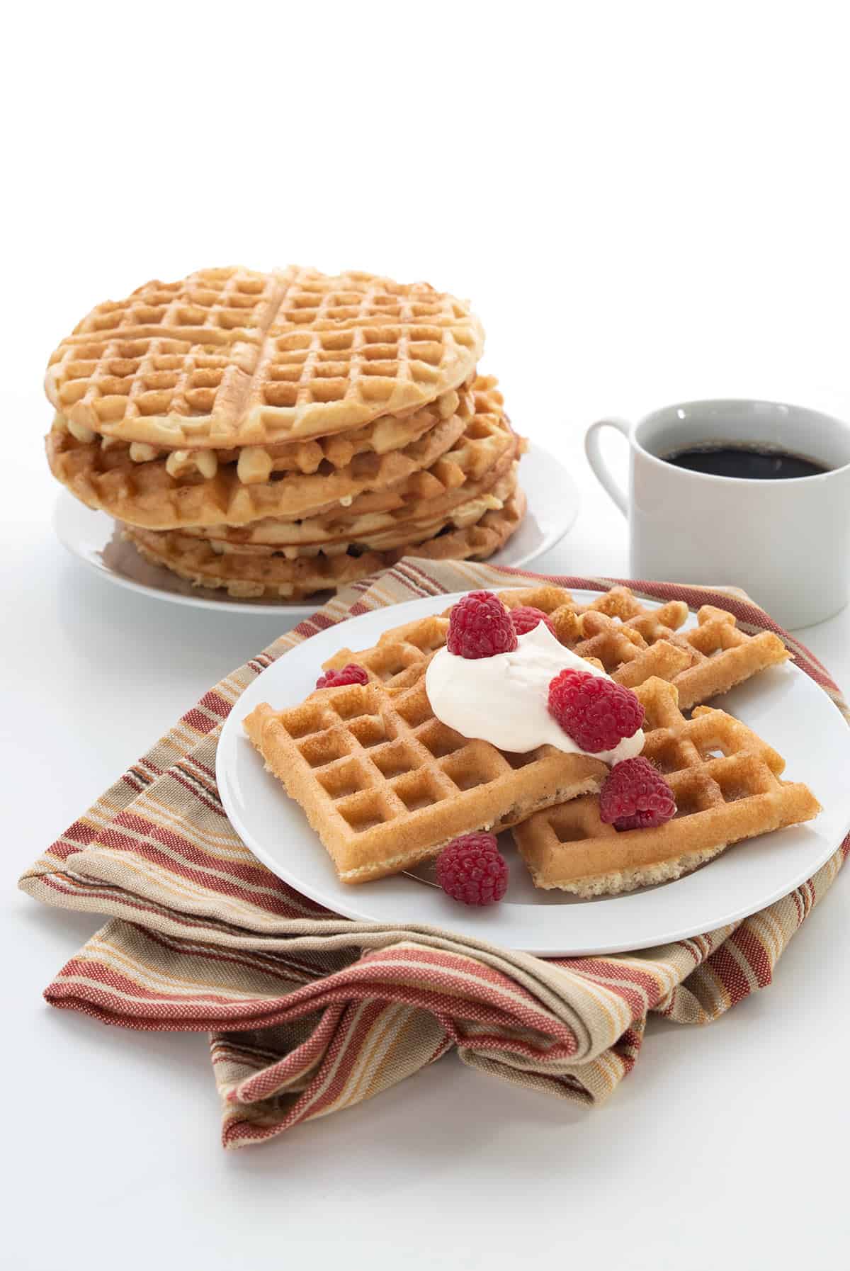 Keto waffles on a white plate over a striped tan and red napkin. A stack of waffles and a cup of coffee in the background.